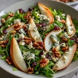 A close-up of an Arugula and Pear Bowl with peppery greens, sweet pears, creamy cheese, and crunchy nuts on a rustic table.