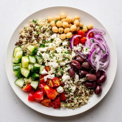 A vibrant Greek Power Salad in a white bowl with quinoa, lentils, chickpeas, tomatoes, cucumber, and feta.