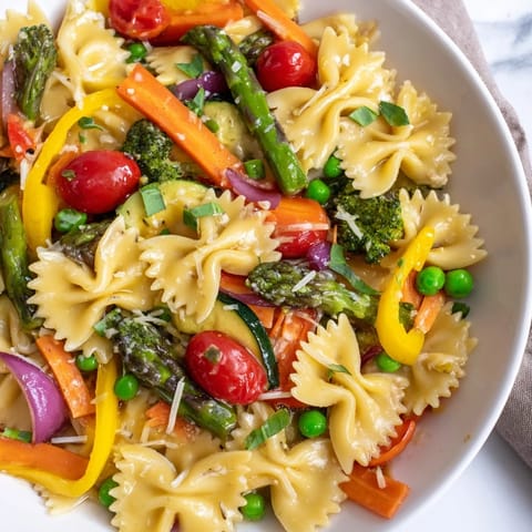A close-up of Rainbow Veggie Pasta Primavera highlights steaming, vibrant veggies and herbs on a white dinner plate.  