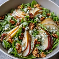 Fresh arugula and sliced pears topped with crumbled blue cheese and toasted walnuts in an Arugula and Pear Bowl, drizzled with balsamic dressing.